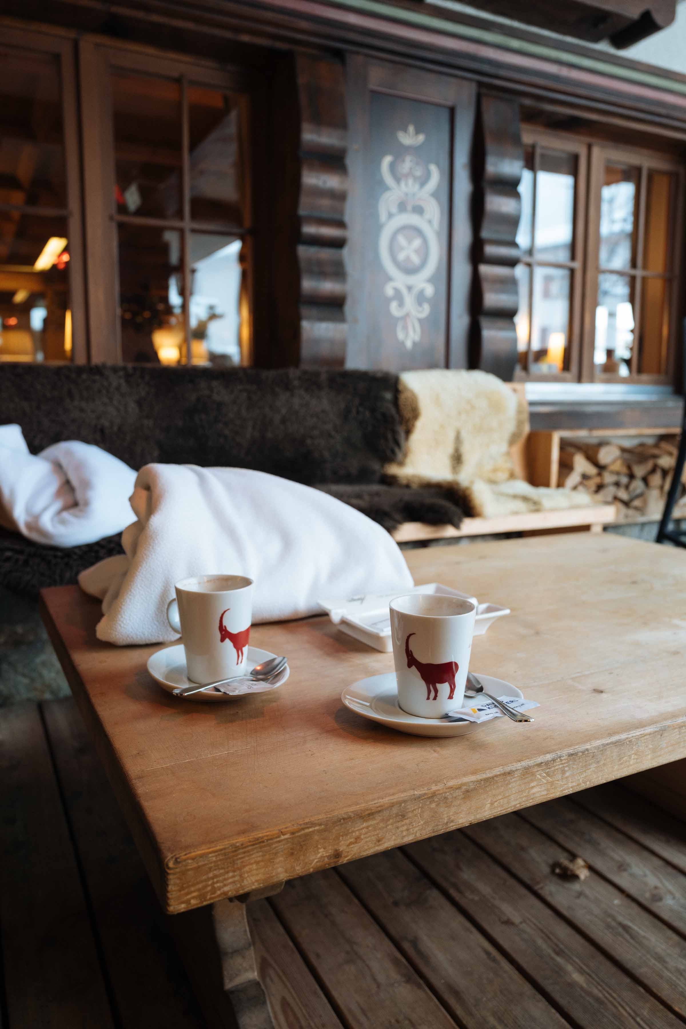 Travel to the alps in winter for a winter wonderland vacation! Cozy up with a blanket at one of these outdoor smoking lounges with heaters and sheep skins to stay warm. Order anything from the bar to enjoy or a hot chocolate while you watch the snow fall and the red Gotschnabahn cable car go up and down the mountain filled with skiers. | thetasteedit.com #travel #switzerland #winter #drink
