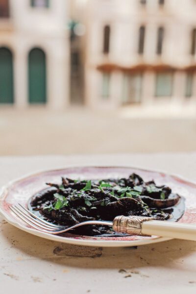 A close-up shot of a plate of Seppie al Nero – tender cuttlefish pieces stewed in their dark ink, garnished with fresh parsley, set against an old Venetian street backdrop.
