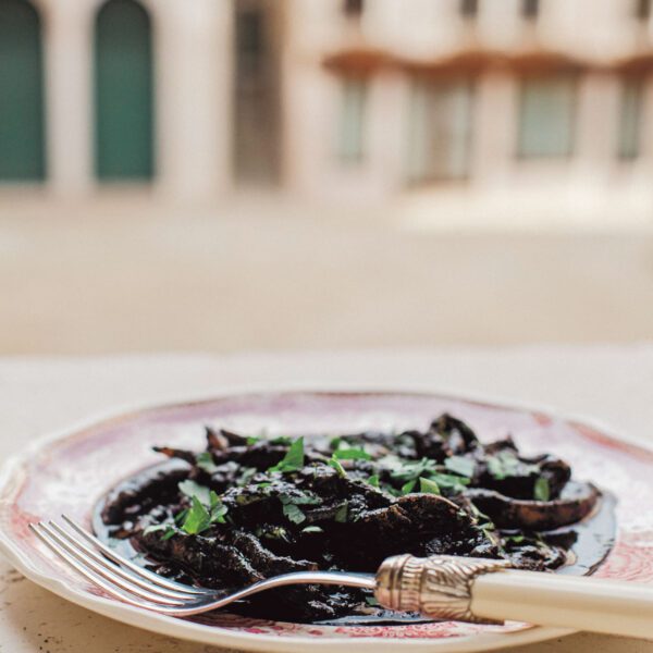 A close-up shot of a plate of Seppie al Nero – tender cuttlefish pieces stewed in their dark ink, garnished with fresh parsley, set against an old Venetian street backdrop.
