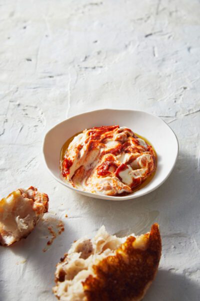 A bowl of creamy red pepper dip swirled with cream cheese, drizzled with olive oil, accompanied by warm toasted bread pieces on a white background.