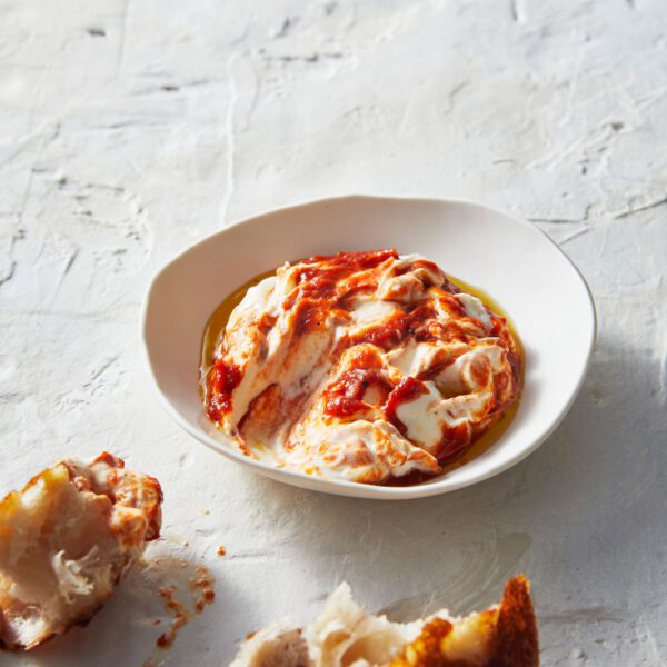 A bowl of creamy red pepper dip swirled with cream cheese, drizzled with olive oil, accompanied by warm toasted bread pieces on a white background.