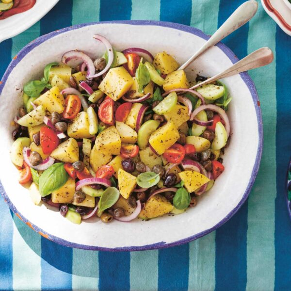 Colorful potato, olive, and caper salad with cherry tomatoes, red onion, cucumber, and fresh basil served in a large bowl on a striped blue tablecloth.