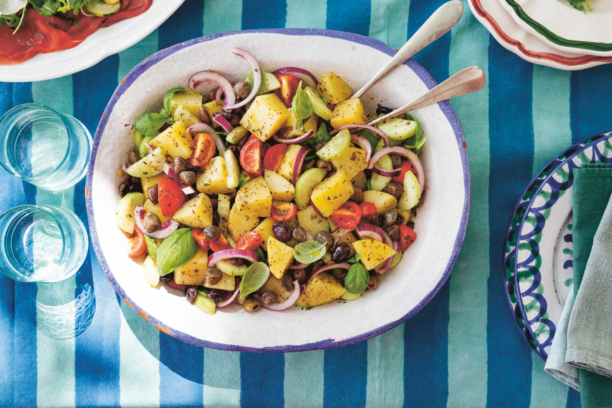 Colorful potato, olive, and caper salad with cherry tomatoes, red onion, cucumber, and fresh basil served in a large bowl on a striped blue tablecloth.