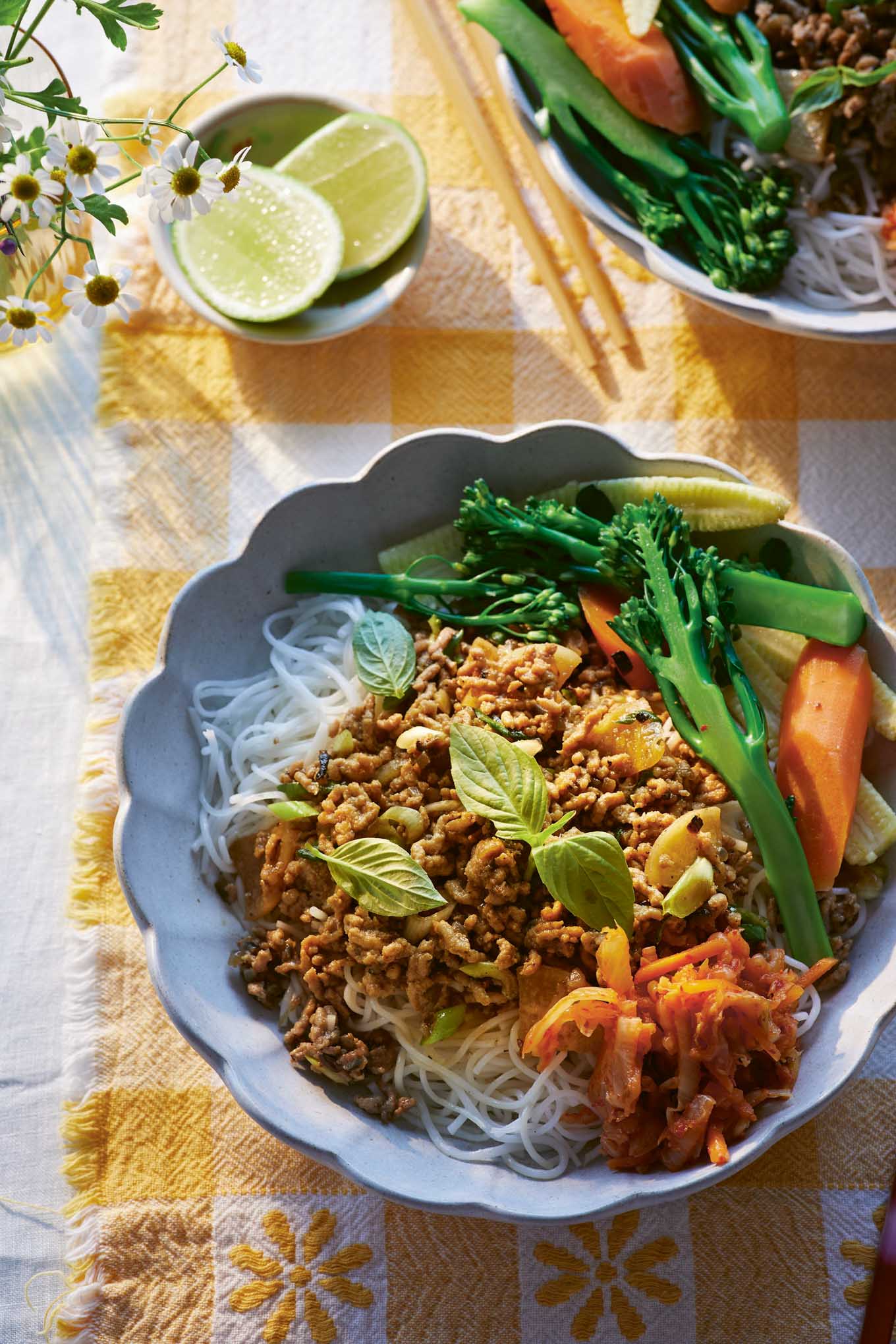 Colorful bowl of Lemongrass Minced Pork Vermicelli with rice noodles, fresh vegetables like broccolini and carrots, kimchi, Thai basil, and lime wedges on a checkered yellow tablecloth