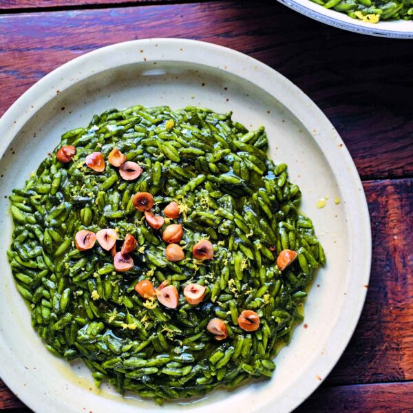 A plate of green orzo pasta, garnished with toasted hazelnuts and lemon zest, sits on a wooden table. Another plate of the same dish is partially visible in the background. by The Taste Edit