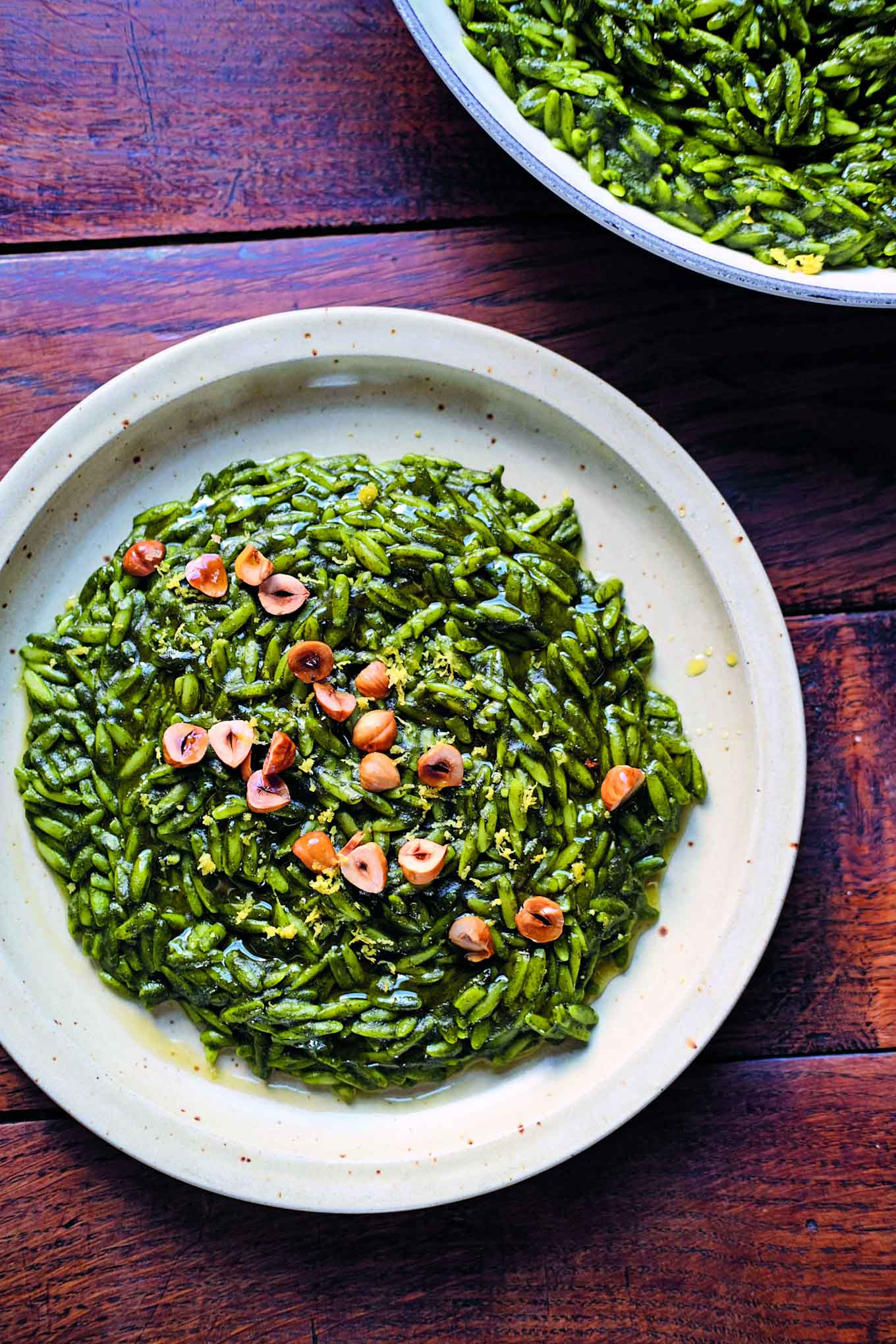 A plate of green orzo pasta, garnished with toasted hazelnuts and lemon zest, sits on a wooden table. Another plate of the same dish is partially visible in the background. by The Taste Edit