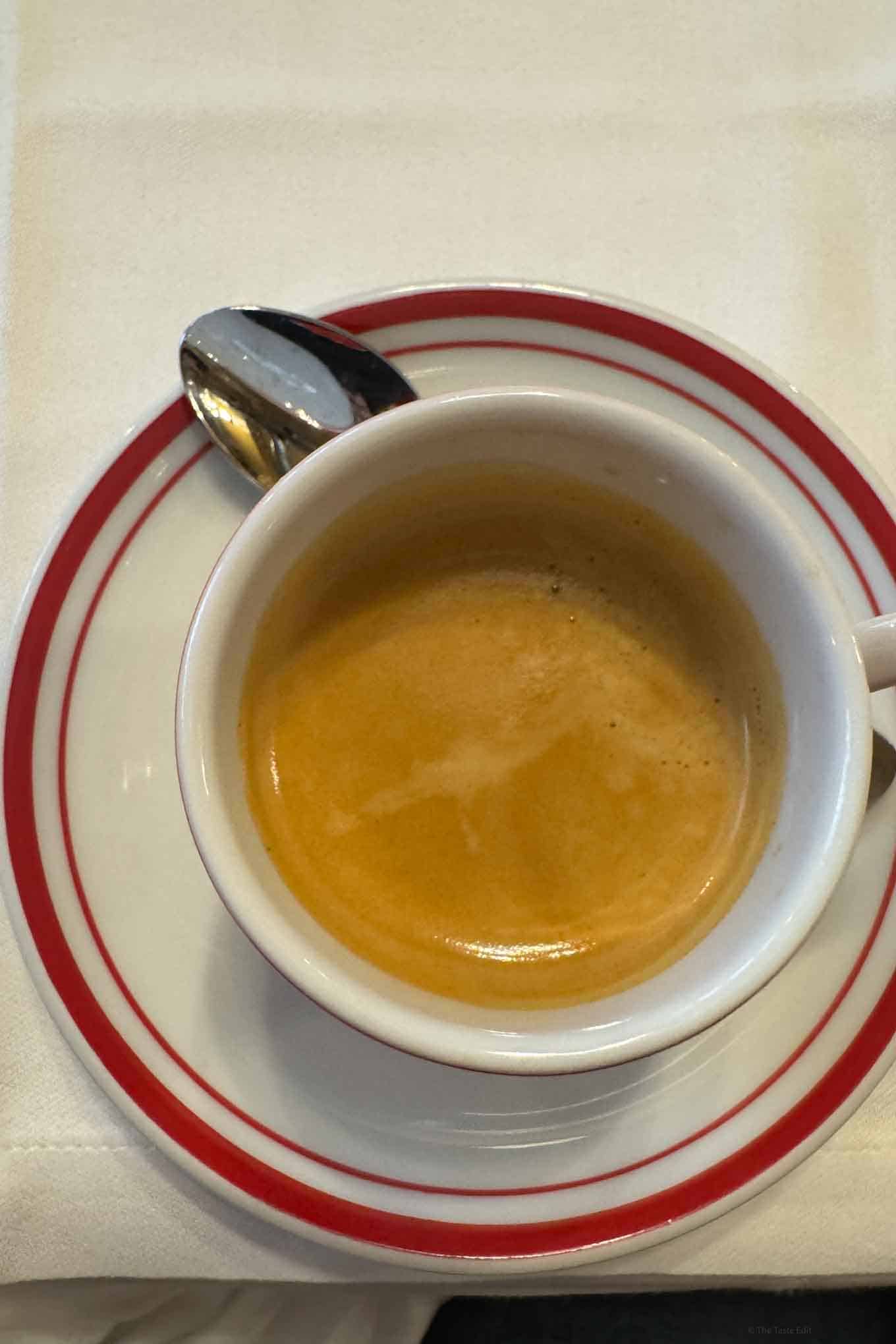 Coffee in a white cup with red accents on a matching saucer, placed on a white tablecloth.