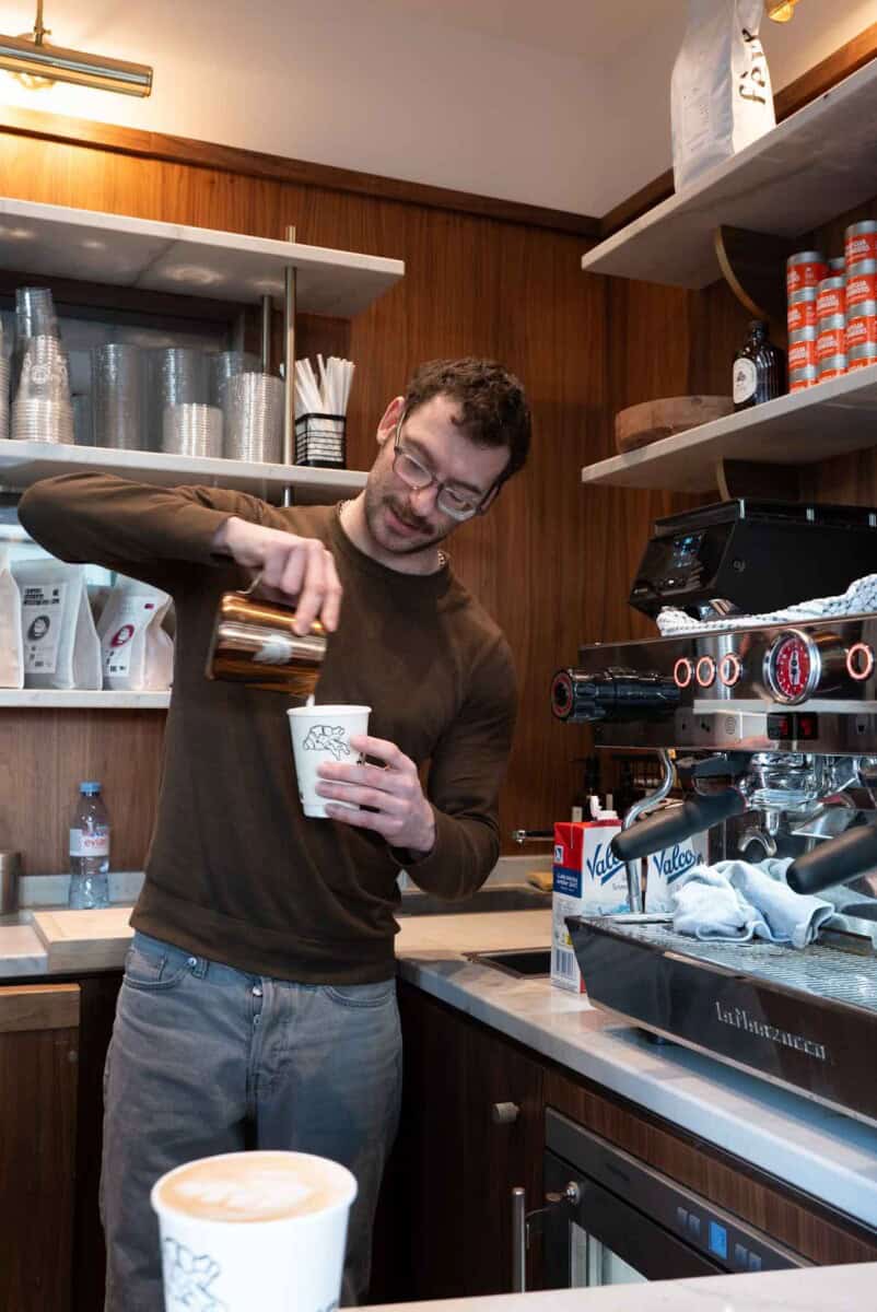 A barista pours steamed milk into a cup of freshly brewed coffee in a warm, inviting café setting.