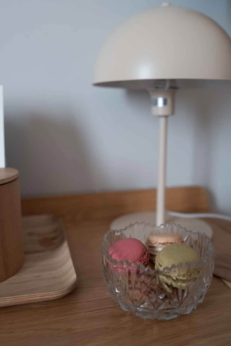 Delicious colorful macarons displayed in a glass bowl on a wooden table, with a modern white lamp in.